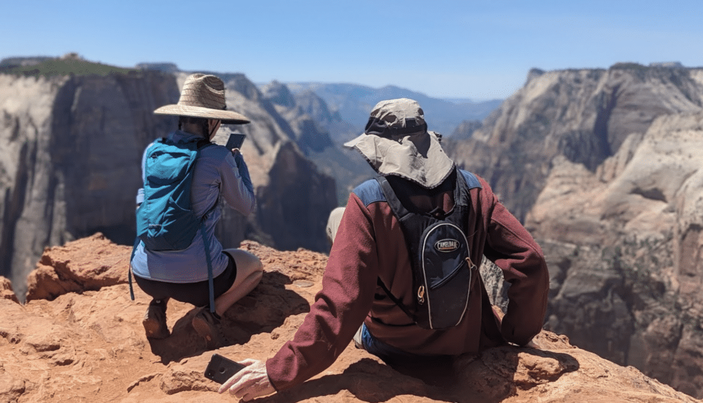 Two hikers sit on a rocky ledge, one photographing the canyon view while the other relaxes.